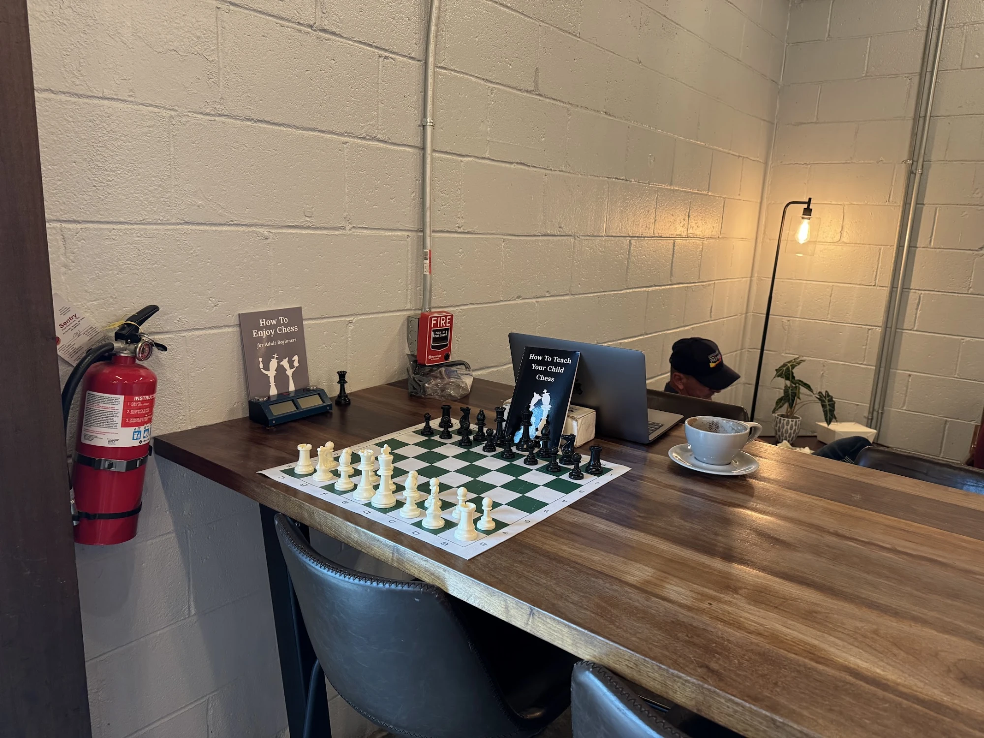 Chess board mid-game with two chess books, a chess clock, a fire extinguisher, a closed laptop, and a coffee saucer on the wooden table.